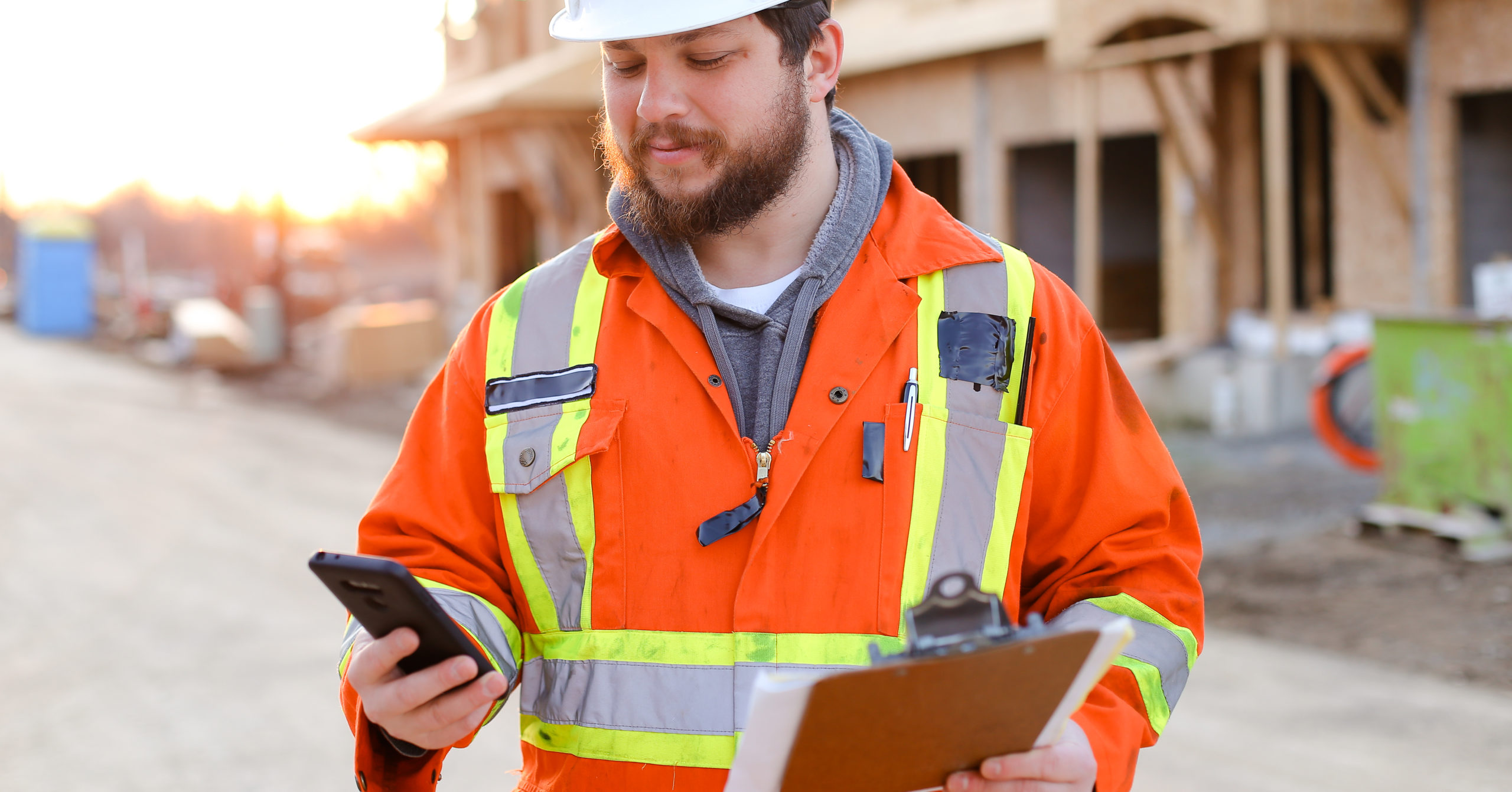 A builder is holding and looking at a smart phone in his right hand while holding a clipboard in his other hand.