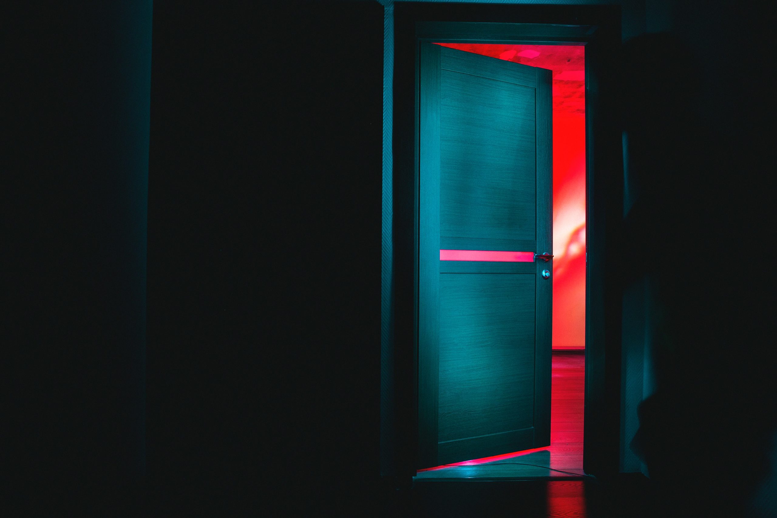 A pale door shadowed in darkness stands ajar, showing an alarmingly red room past it.